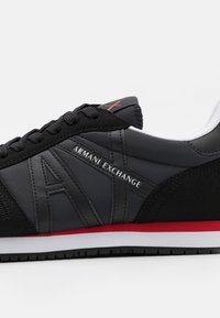 Fekete sportcipő sima szintetikus felsőrésszel és bőrhatású díszítéssel. Piros talppal, fűzős záródással és "Armani Exchange" márkajelzéssel rendelkezik.
