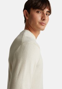 Helles beige Sweatshirt mit langen Ärmeln, geripptem Halsausschnitt und strukturiertem Stoff. Die Passform ist körpernah und betont die Schulternähte.