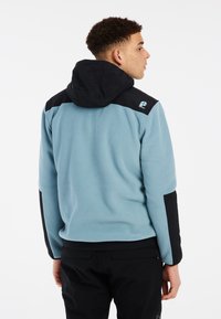 Lichtblauwe fleecejack met een zwarte capuchon en schouderaccenten. Voorzien van een logo op de rechter schouder en zwarte mouwen. Zachte textuur.