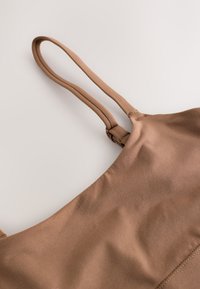 Nahaufnahme eines braunen Stoffkleidungsstücks mit dünnen, verstellbaren Schulterriemen vor hellem Hintergrund.