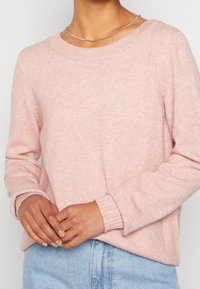 Ljusrosa stickad tröja med vid halsringning, ribbade ärmslut och avslappnad passform, i kombination med ljusblå denim. Händerna är placerade fram.