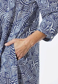 Marineblau und weiß gemustertes Kleid mit geometrischen Formen. Lange Ärmel, eine Tasche auf der linken Seite und ein zartes Goldarmband am Handgelenk.