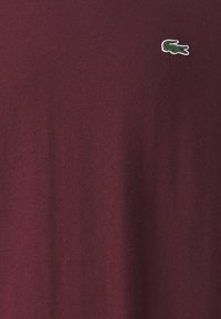 Burgundska pletena jopica s majhnim zelenim veznim logotipom krokodila. Tkanina ima gladko teksturo in klasičen okrogli izrez.