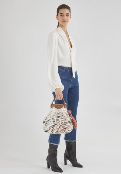 Weiße Bluse mit langen Ärmeln und Bindegürtel, kombiniert mit blauen Jeans und schwarzen kniehohen Stiefeln. Das Model hält eine cremefarbene Netztasche mit braunen Akzenten.