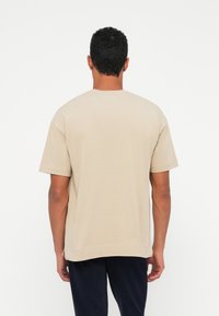 Beige t-paita lyhythihaisilla, pyöreällä kauluksella ja väljässä mitassa, valmistettu pehmeästä puuvillasta. Ei näkyviä kuvioita tai logoja. Sivusaumareiät helmassa.