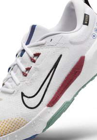 Baskets blanches Nike avec logo noir, accents rouges et bleus, gouttes d'eau à la surface et étiquette imperméable Gore-Tex au niveau du talon.