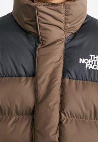 Chaqueta acolchada en marrón y negro, con un cuello alto, diseño acolchado y un logo prominente en el pecho. Textura suave y material sintético.