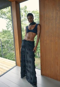 Marineblauwe twee-delige outfit bestaande uit een cropped top en lange rok met textuur en bloemborduursels, geplaatst in een houten interieur met groenvoorziening buiten.