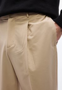 Μπεζ υφασμάτινο παντελόνι με πιέτες και τσέπες, φορεμένο με μαύρο ριπ πουλόβερ, το δεξί χέρι τοποθετημένο μέσα στην τσέπη.