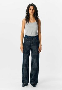 Pantalons larges en denim foncé avec un motif géométrique, taille haute, deux poches avant, associés à un débardeur gris. Cheveux bouclés.