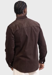 Camisa de pana marrón con yugo trasero y mangas largas, que presenta puños abotonados y una textura suave. Acentuada con botones blancos.