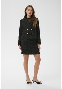 Juodas tweedo kostiumas su dvieiliai sagstoma švarku ir prigludusia suknele, puoštas auksinėmis sagomis. Modelis dėvi juodus aukštakulnius, šviesūs plaukai laisvai krinta.