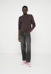 Νεαρός άνδρας που φοράει σκούρο καφέ ριμπ κορμάκι, χαλαρά μαύρα τζιν και ροζ αθλητικά παπούτσια, στέκεται μπροστά σε λευκό φόντο με το ένα χέρι στην τσέπη.