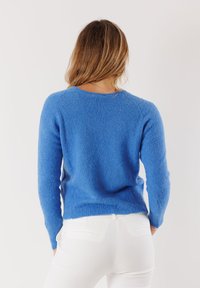 Maglione blu lavorato a maglia con maniche lunghe, scollo rotondo e tessuto testurizzato. Indossato con pantaloni bianchi, che mostrano una silhouette aderente.