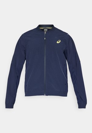 Dunkelblaues Zip-Up-Jackett mit Rundhalsausschnitt, elastischen Bündchen und perforierten Details auf den Schultern. Logo-Akzent in Gelb. Leichtes Material.