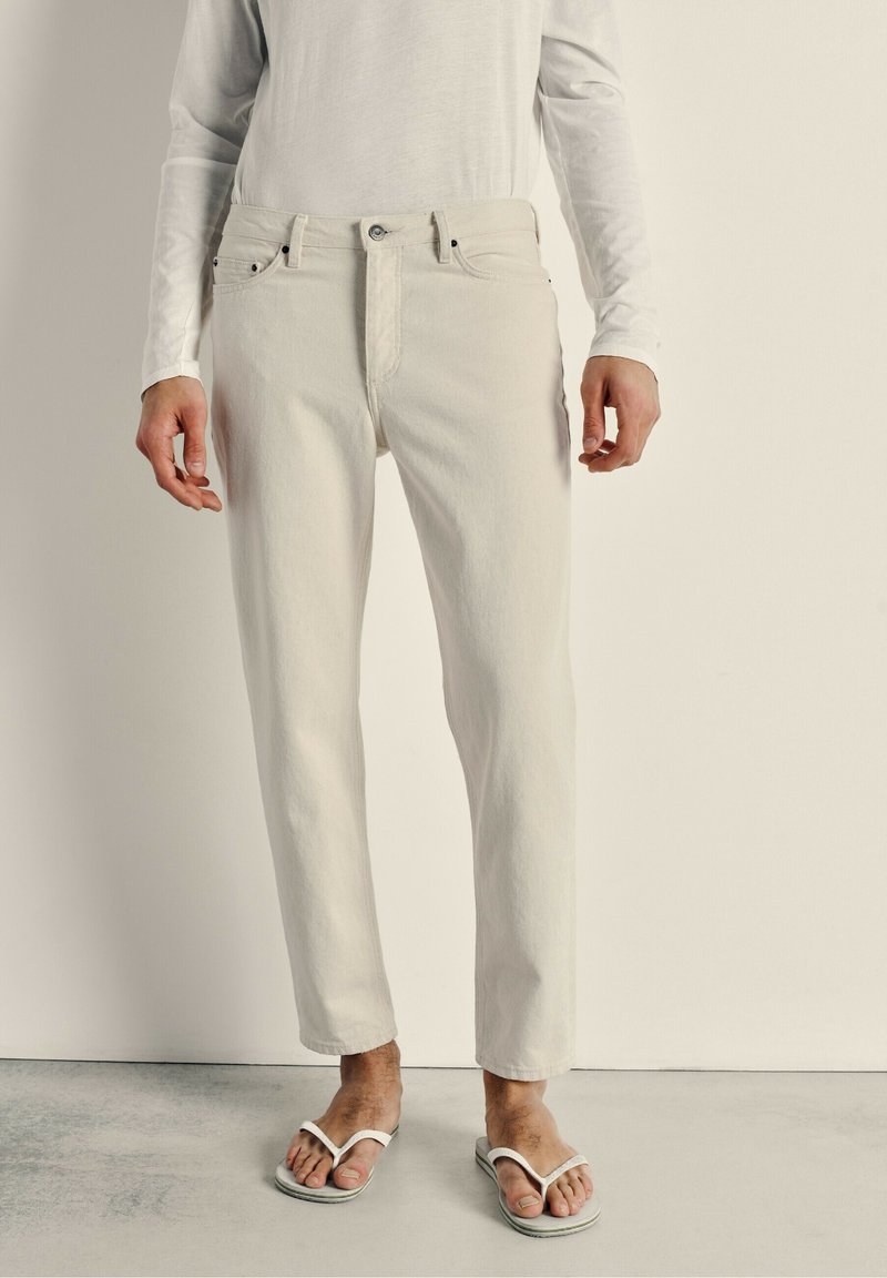 Jean en denim beige clair avec une coupe droite, cinq poches et une fermeture à bouton sur le devant. Associé à des tongs blanches et une chemise à manches longues.