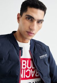 Jūrinis bombonešis su užtrauktuku, gumuotu apykakle ir „Red Bull Racing“ logotipu; dėvimas virš balto megztinio su raudonais blokiniais rašmenimis, užrašytomis „RACING“. 