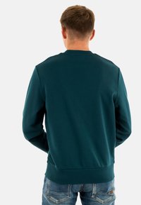 Felpa teal scuro con scollo rotondo e polsini a coste, abbinata a jeans in denim chiaro, mostrando una vista posteriore semplice.