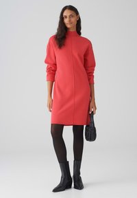 Abito in maglia corallo con collo alto, spalle scese, maniche lunghe e una silhouette dritta. Indossato con collant neri e stivaletti alla caviglia.