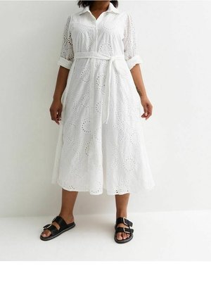 Vrouw draagt een witte midi-jurk met eyeletdetails, opgerolde mouwen en een ceintuur, gecombineerd met zwarte sandalen met twee gespen.
