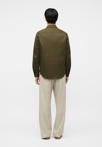 Άνδρας που στέκεται με την πλάτη στραμμένη, φοράει ένα ελαιδί μακρυμάνικο πουκάμισο και μπεζ παντελόνι φαρδιάς γραμμής σε απλό λευκό φόντο.