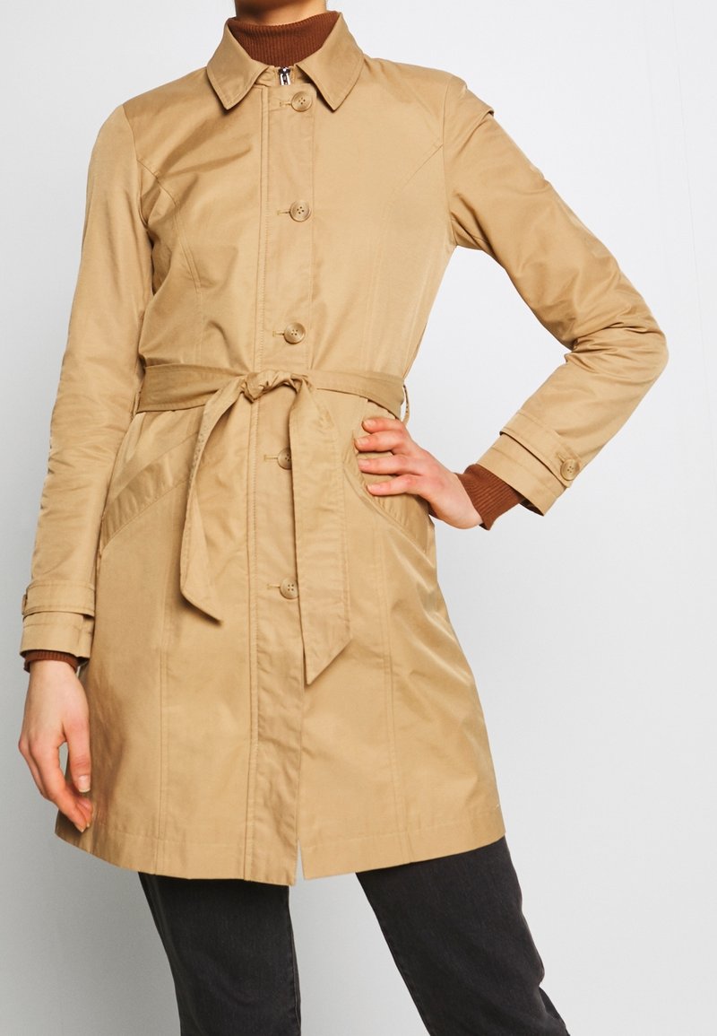 Beige, vyötetty trench-takki suoran siluetin kanssa, nappisuljenta, lovettu kaulus ja kaksi sivutaskua. Sujuva rakenne, kevyet materiaalit.