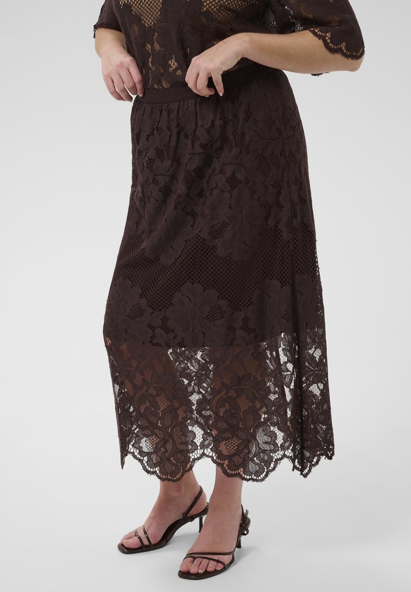Femme portant une jupe midi en dentelle marron foncé transparente et un top en dentelle assorti, associée à des sandales à talons hauts à fines brides, debout sur un fond uni.
