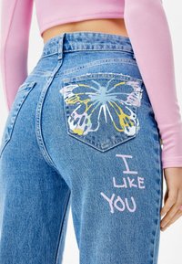 Ljusblå denimjeans med en fjärilsdesign i pastellfärger på bakfickan och texten "JAG GILLAR DIG" i rosa på benet.