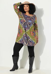 Femme portant une tunique à motifs multicolores, des leggings noirs, des bottes de cowboy noires et un chapeau beige, posant avec un bras levé.