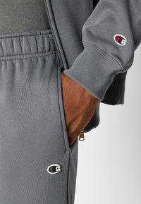 Grijze sweatshirt en joggingbroek gemaakt van zacht materiaal. Voorzien van ribbelmanchetten, een elastische tailleband en een logo-opdruk op de linkerborst en de broek.