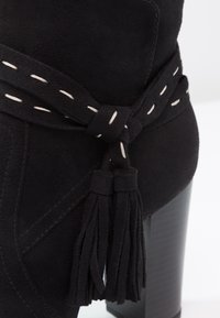 Bottine en daim noir avec une bride décorative cousue et des pompons, dotée d’un talon bloc empilé.