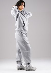 Young woman in gray tracksuit adjusting hoodie, standing sideways against plain gray background, wearing white socks and gray sneakers.