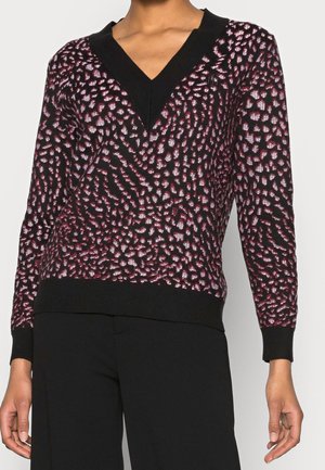 Frau trägt einen schwarz-rosa gemusterten V-Ausschnitt-Pullover kombiniert mit schwarzen Hosen und steht vor einem schlichten hellen Hintergrund.