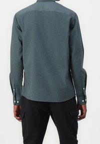 Uomo che indossa una camicia a maniche lunghe di un verde scuro con polsini con bottoni e pantaloni neri con tasche a cargo, visto da dietro.