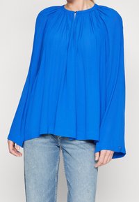 Blusa blu con maniche ampie, scollo arricciato e tessuto liscio. Abbinata a jeans azzurri chiari. La blusa presenta una leggera lucentezza.