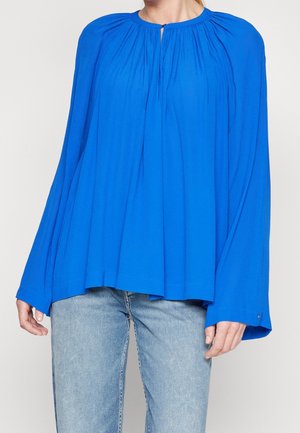 Blaue Bluse mit weiten Ärmeln, gerafftem Ausschnitt und glattem Stoff. Kombiniert mit hellblauen Jeans. Die Bluse hat einen dezenten Glanz.