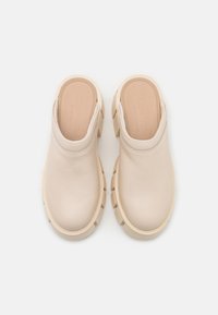 Beżowe klapki wsuwane z grubą podeszwą i gładkimi skórzanymi cholewkami, widok z góry na białym tle.