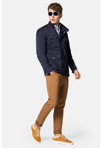 Granatowa ocieplana kurtka z czterema przednimi kieszeniami, zestawiona z sandałami w kolorze brązowym o dopasowanym kroju i żółtymi trampkami. Model nosi okulary przeciwsłoneczne.