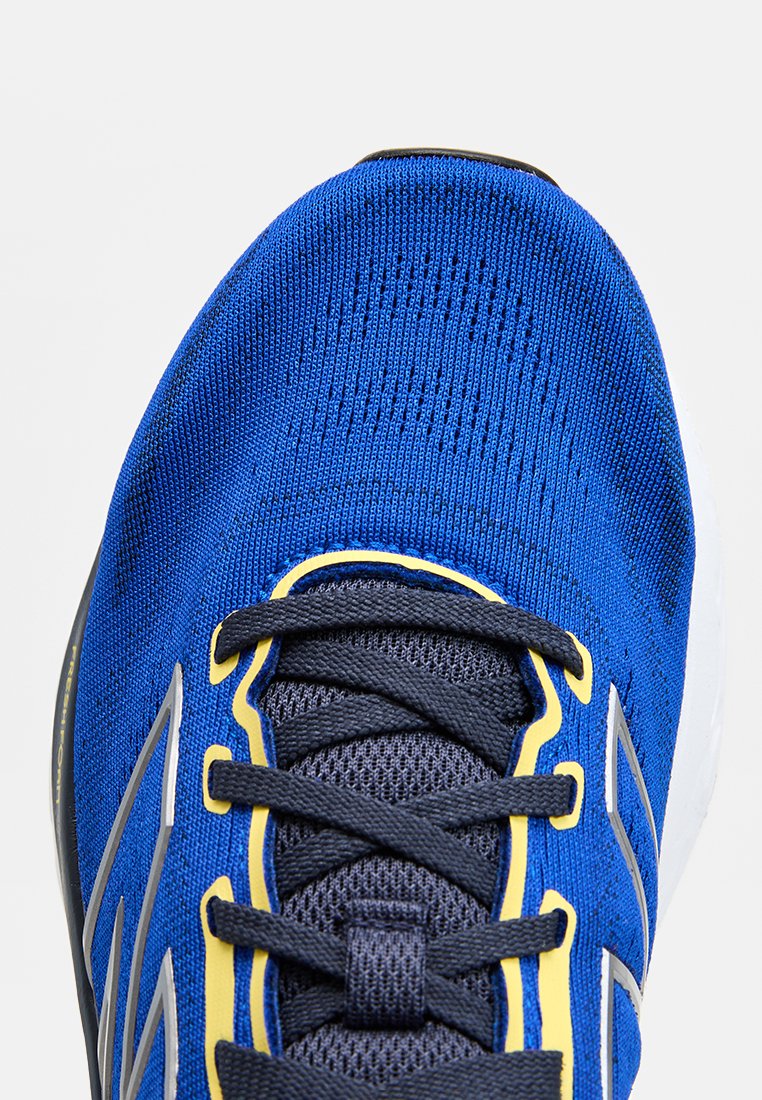 Zapatilla deportiva azul con textura de punto, con cordones gris oscuro, acentos amarillos a lo largo de los lados y marca reflectante en plata.