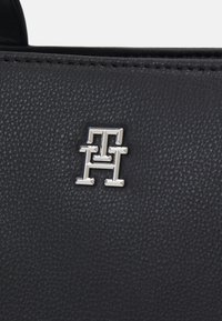 Primo piano di una borsa a tracolla nera in pelle con superficie testurizzata e un logo metallico argentato, che presenta un design geometrico con angoli acuti.