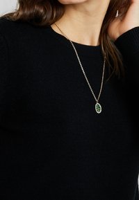 Goldkette mit ovalem Anhänger, der grüne Akzente und einen strukturierten Hintergrund aufweist, getragen über einem schwarzen Pullover.