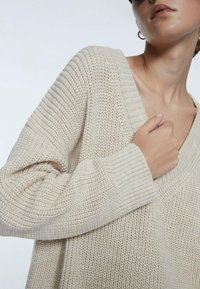 Beige gebreid sweater met een V-hals, voorzien van geribbelde manchetten en zoom. De textuur is los, met een chunky, gelaagd ontwerp.