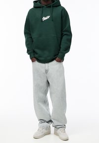 Sweat à capuche vert avec logo blanc sur la poitrine, associé à un jean baggy gris clair et des baskets claires. Coupe décontractée avec des poignets élastiques.