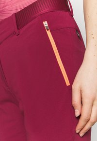 Pantalon bordeaux avec une poche zippée orange. Confectionné dans un tissu lisse et léger, doté d'une ceinture texturée. Gros plan sur une vue latérale.