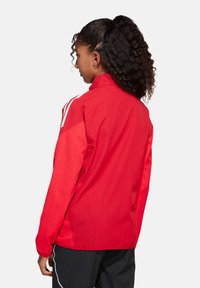 Rode jas met een staande kraag, witte accenten op de schouders, lichtgewicht stof en een gladde textuur, ontworpen voor sportkleding.
