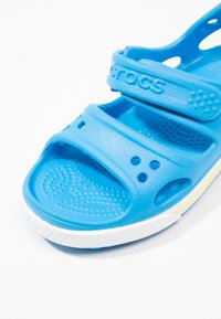 Sininen vaahtomuovisandaali, jossa on kaksi säädettävää hihnaa, rei'itetty muotoilu hengittävyyden vuoksi, valkoinen pohja keltaisella korostuksella, teksturoitu pohja pitävyyden parantamiseksi.