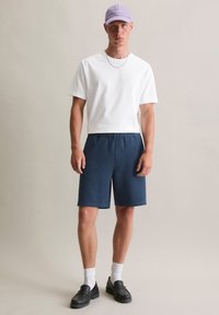 Witte korte mouwen t-shirt, marineblauwe wafel-gehaakte shorts, paarse pet, zwarte instappers en witte sokken. Eenvoudige, casual outfit met een relaxte pasvorm.