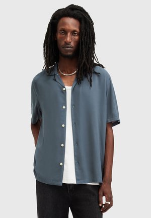 Chemise à manches courtes à boutons en tissu bleu ardoise doux, avec une coupe décontractée, un col à encoches et des boutons blancs, portée par-dessus un T-shirt blanc.