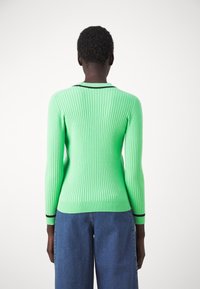 Personne aux cheveux courts portant un pull vert néon côtelé ajusté avec des bordures noires et un jean bleu ample, de dos sur un fond blanc.