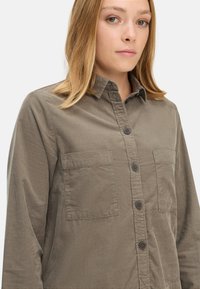 Chemise en velours côtelé kaki avec un devant à boutons, deux poches poitrine et un col. Texture douce et coupe décontractée.